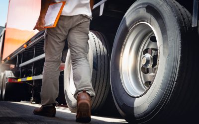 Man walking alongside a truck, holding a clipboard, inspecting large truck tires, emphasizing truck maintenance and safety.